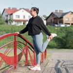 A woman in active wear performs a stretching exercise on a red bridge in a suburban park setting.